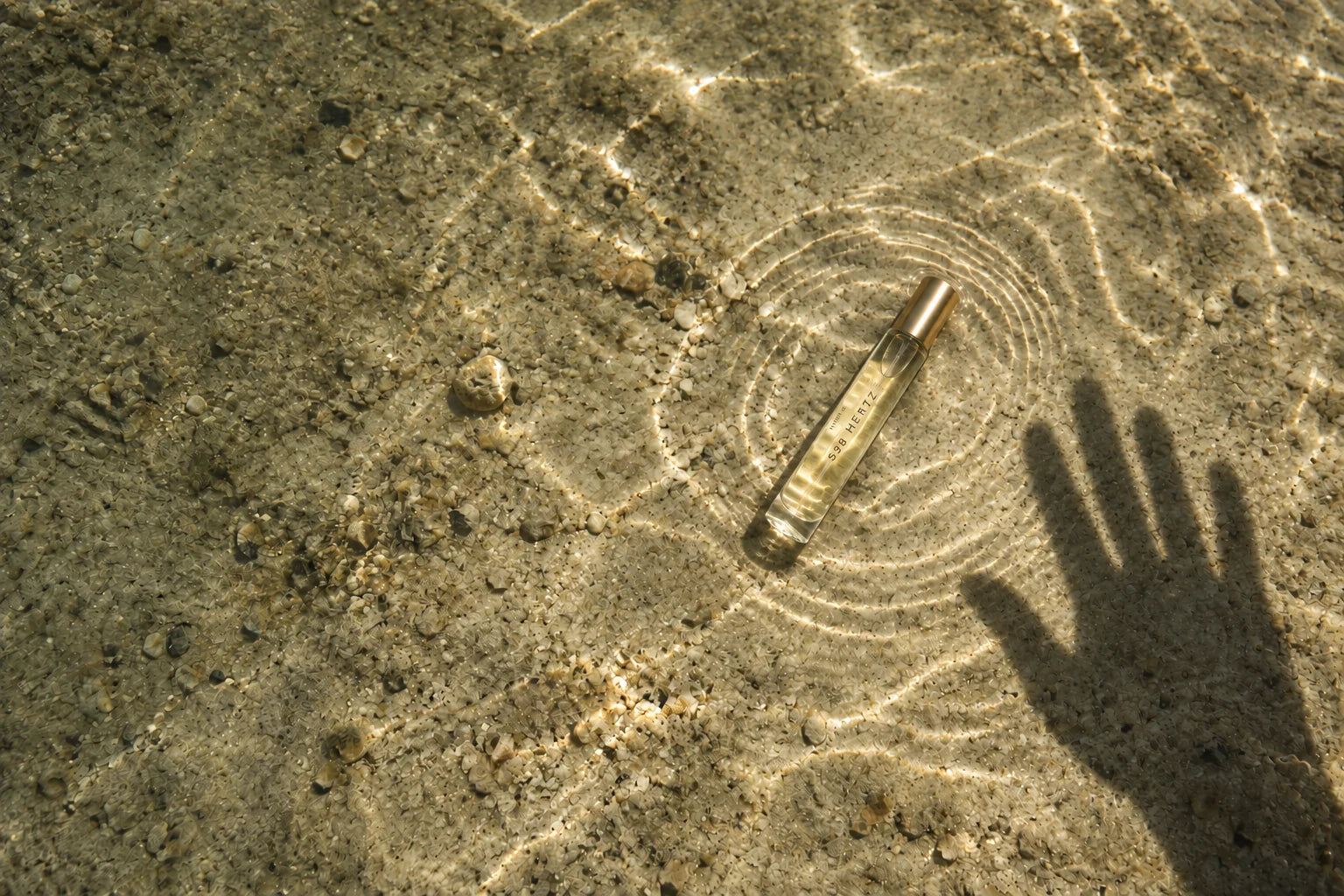 A clear gold-capped 528 Hertz bottle lies over sand beneath shallow rippling water beside a hand shadow.