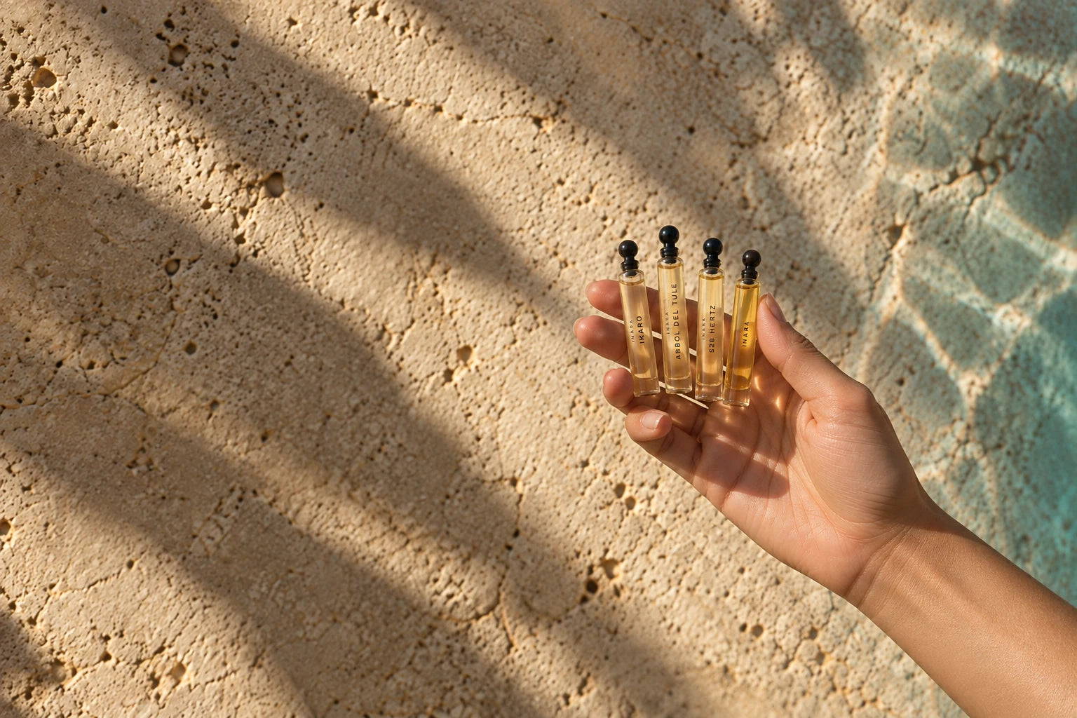 A hand holds four small clear fragrance bottles with black rounded stoppers against sunlit textured stone.